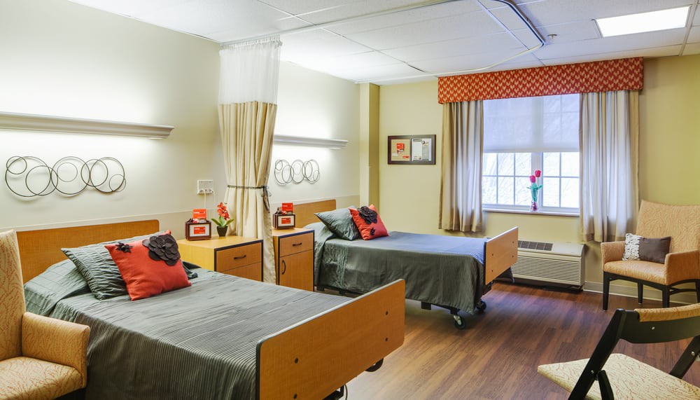 A hospital room with two beds, separated by a privacy curtain, featuring decorative pillows and a large window.