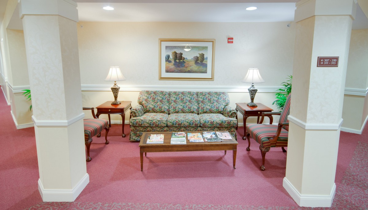A cozy living room with a floral sofa, chairs, and wooden tables, complete with lamps and a painting on the wall.