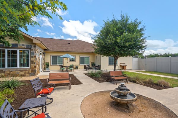 A beautiful backyard with a patio, seating area, and a small fountain surrounded by greenery on a sunny day.