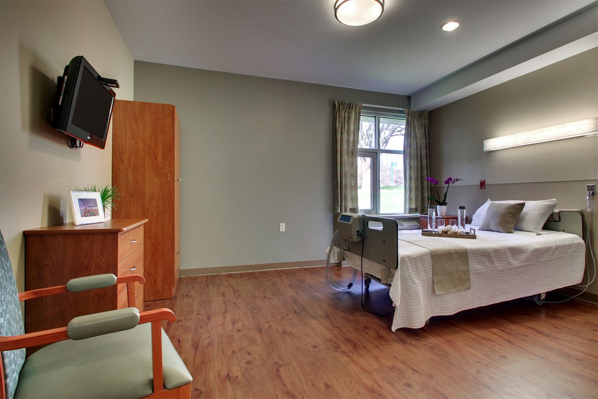 A hospital room with a bed, chair, television, and large window with curtains.