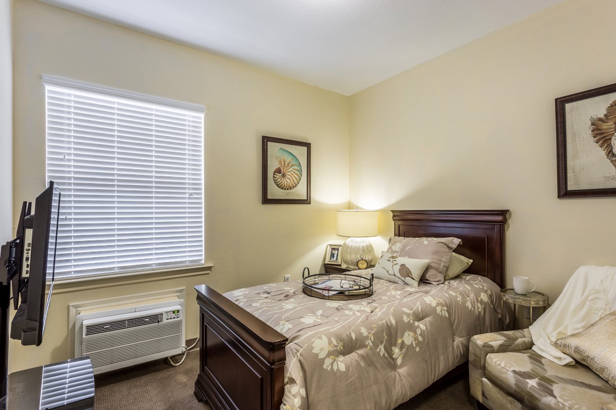 A cozy bedroom with a single bed, adorned with floral bedding, a bedside lamp, and an upholstered chair by the window.