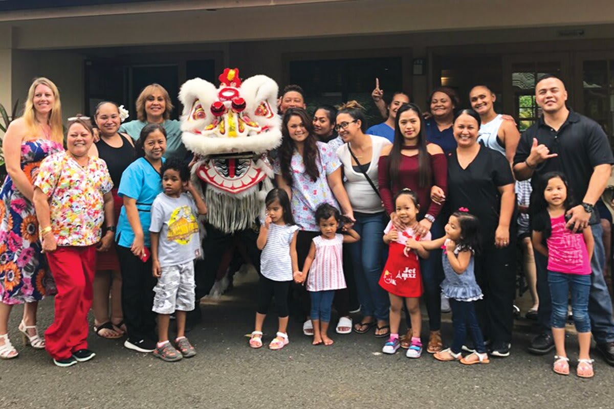 A diverse group of people, including children and adults, gather around a lion dance costume during an outdoor celebration.