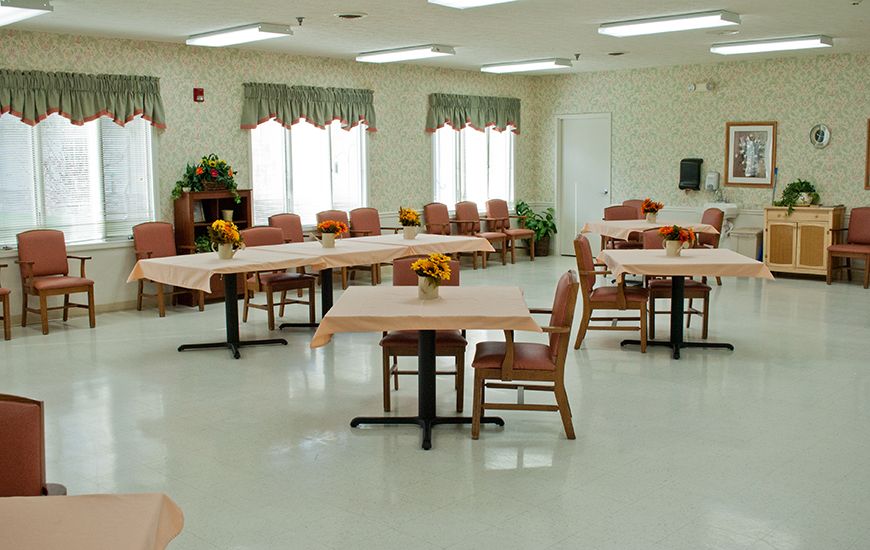An empty dining room with tables and chairs arranged neatly, featuring floral decorations and large windows with curtains.