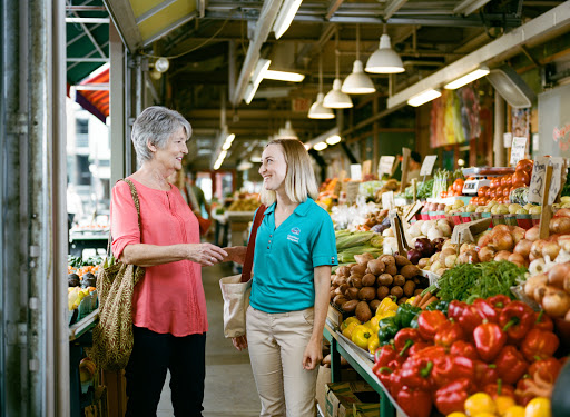 Two women engage in conversation at a vibrant market, surrounded by fresh fruits and vegetables.