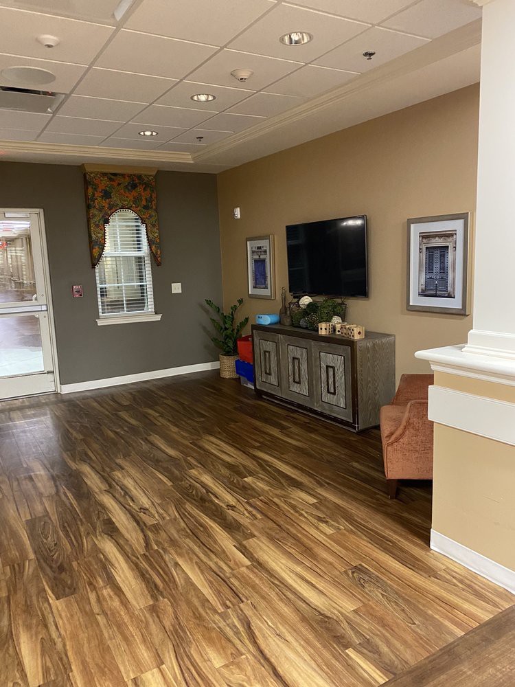 A cozy living room with a hardwood floor, featuring a television, cabinet, plants, and large windows.