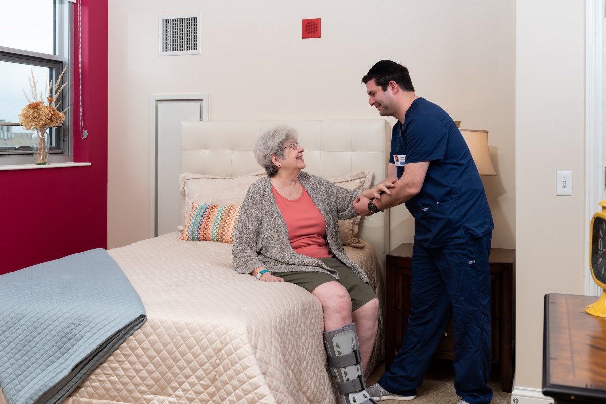 A caregiver assists an elderly person in a bedroom setting.