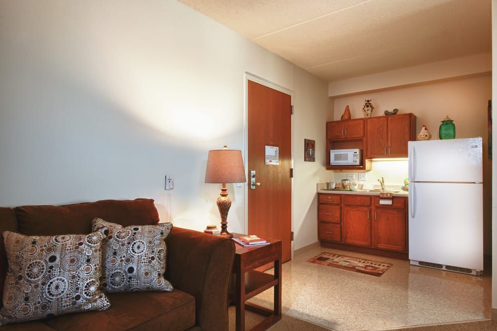 A cozy apartment interior featuring a comfortable living area with a sofa and lamp, adjacent to a compact kitchenette with wooden cabinets.