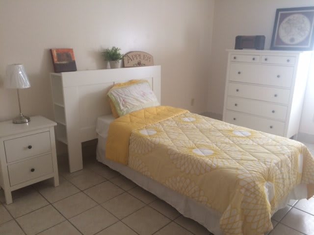 A minimalist bedroom with a single bed featuring yellow bedding, accompanied by white furniture including a nightstand and dresser.