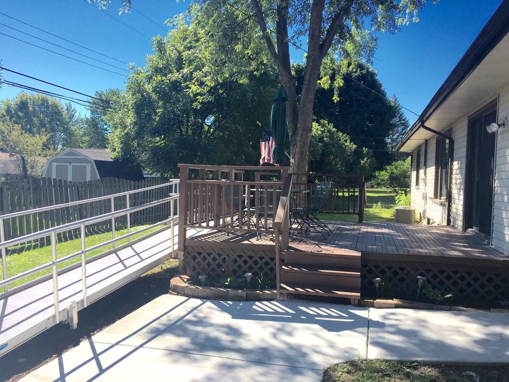 A sunny backyard scene with a wooden deck, accessible ramp, and lush green trees.