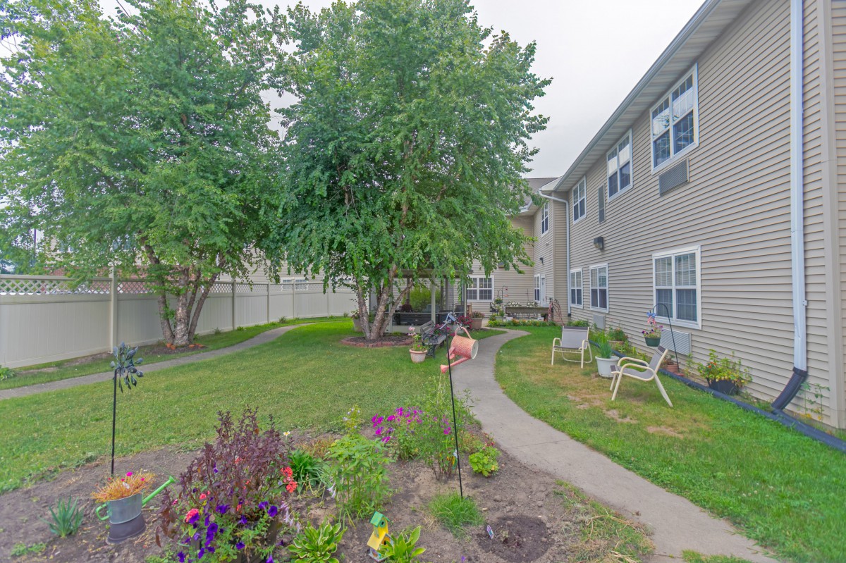 A serene garden with a pathway surrounded by greenery and flowers, adjacent to residential buildings.