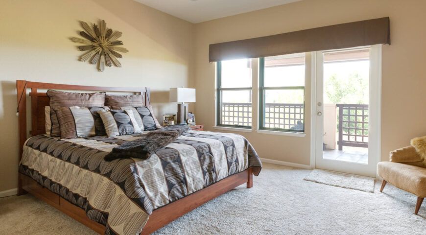 A well-lit bedroom featuring a neatly made bed with patterned bedding, large windows, and stylish decor.