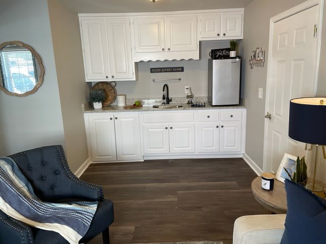 A cozy kitchenette with white cabinets and a small fridge, featuring minimalist decor and wood flooring.