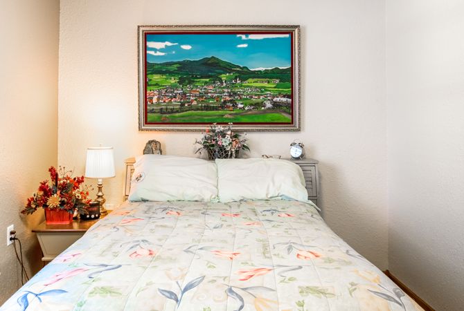 A cozy bedroom with a bed adorned with floral bedding, accompanied by bedside lamps and a vibrant landscape painting on the wall.