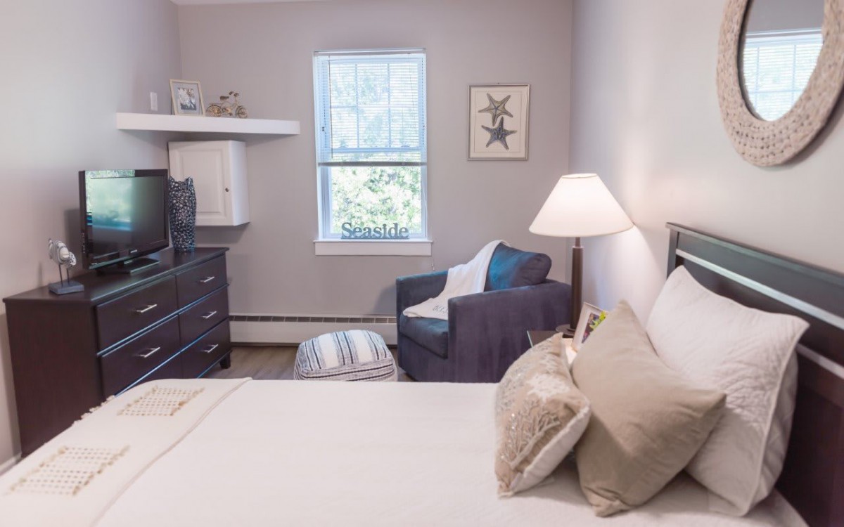 A cozy bedroom with a neatly made bed, an armchair by the window, and soft lighting from a bedside lamp.