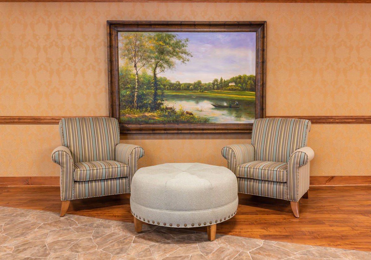 A cozy seating arrangement with two striped armchairs and a round ottoman in front of a landscape painting.