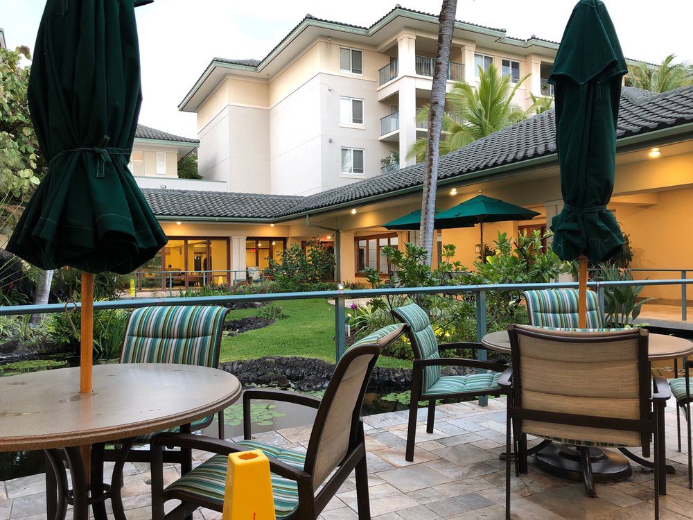 Outdoor patio area with tables, chairs, and umbrellas in a landscaped setting near a multi-story building.