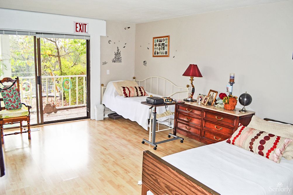 A nursing home bedroom with two beds, a dresser, and a balcony view.