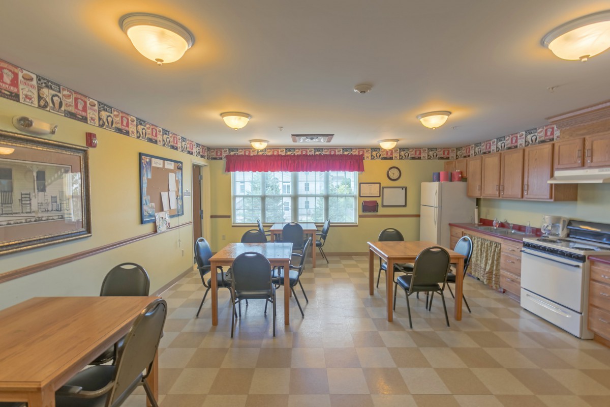 A cozy dining room with several tables and chairs, featuring a small kitchen area and large windows letting in natural light.