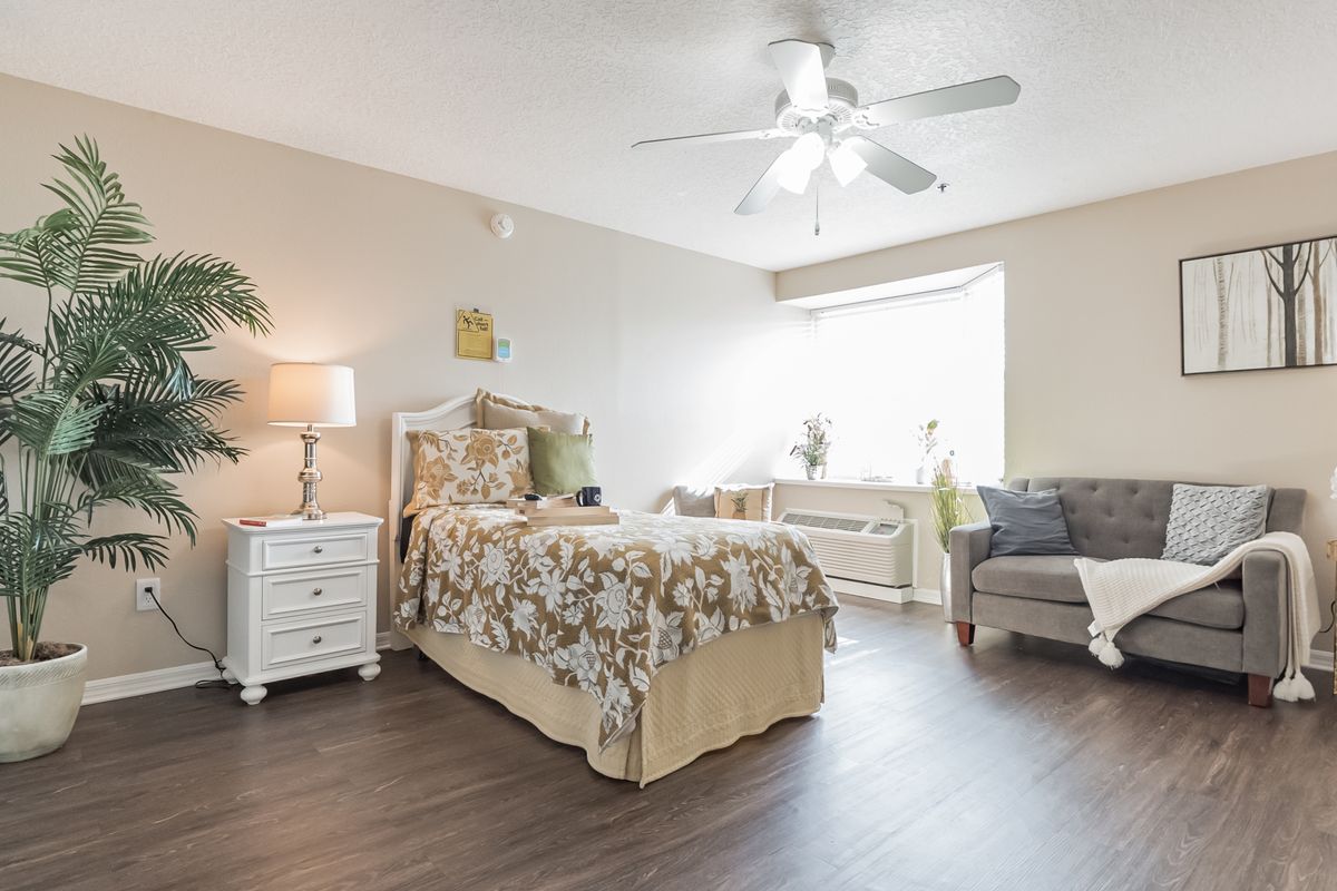A cozy and well-lit bedroom with a floral-patterned bedspread, a bedside table with a lamp, and a comfortable chair by the window.