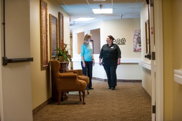 Two people walking and conversing in a hallway with artwork on the walls and furniture along the side.