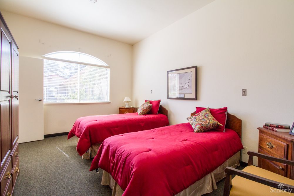 A cozy bedroom with two twin beds featuring bright red bedding, a window allowing natural light, and simple decor.