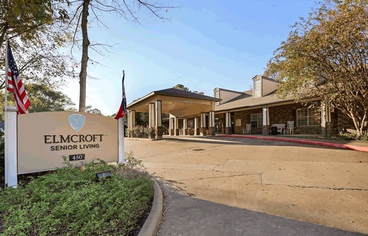 An exterior view of a building with a sign and driveway surrounded by trees under a clear sky.