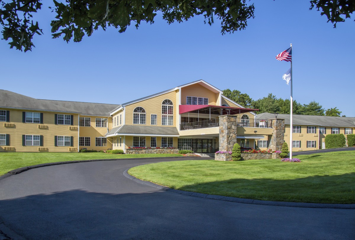 A sunny day view of a residential building with flags in front, surrounded by a well-maintained lawn and driveway.