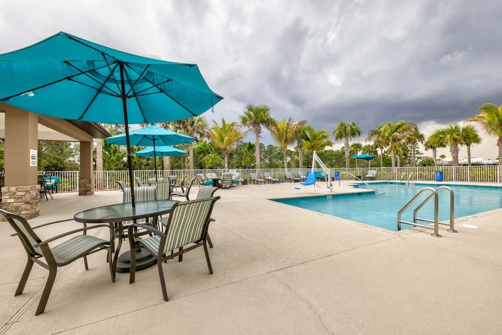 A serene outdoor swimming pool area with tables and umbrellas, surrounded by palm trees under a cloudy sky.