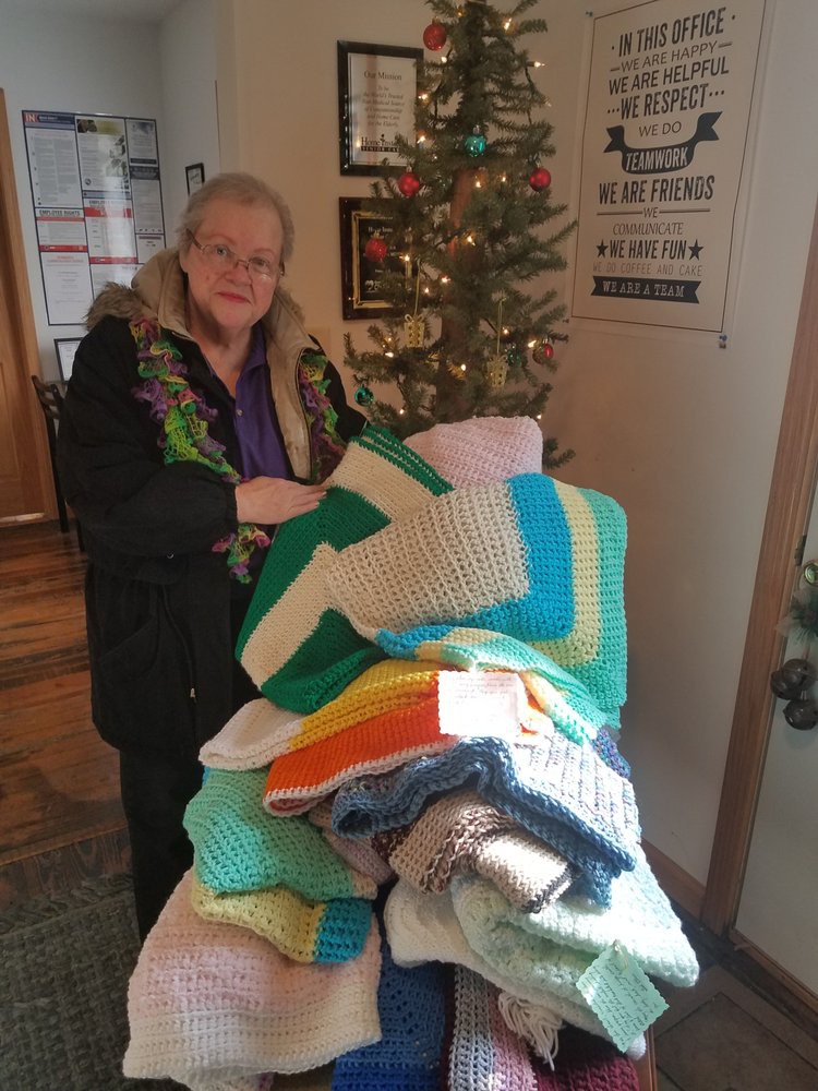 A woman standing indoors holding a collection of colorful crocheted blankets, with a tree in the background.