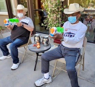 Two people wearing cowboy hats and face masks are seated outside, each holding a toy.