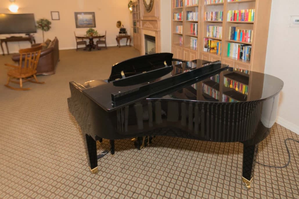 A grand piano is placed in a cozy library room with bookshelves and comfortable seating.