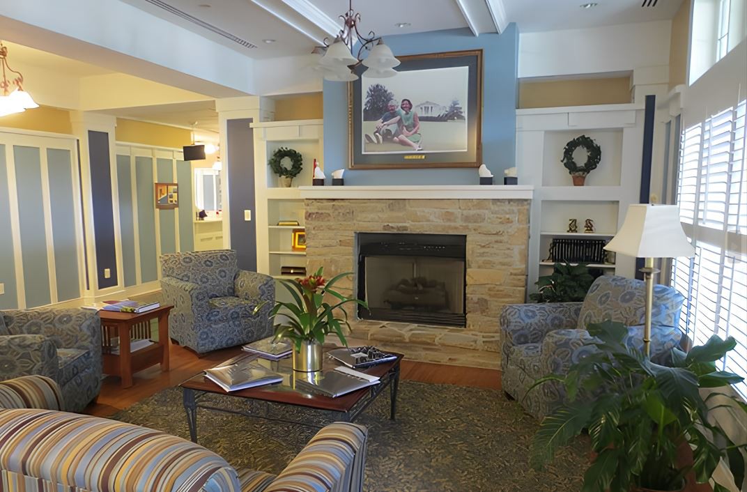 A cozy living room with a central fireplace, surrounded by comfortable seating and decorated with paintings and plants.