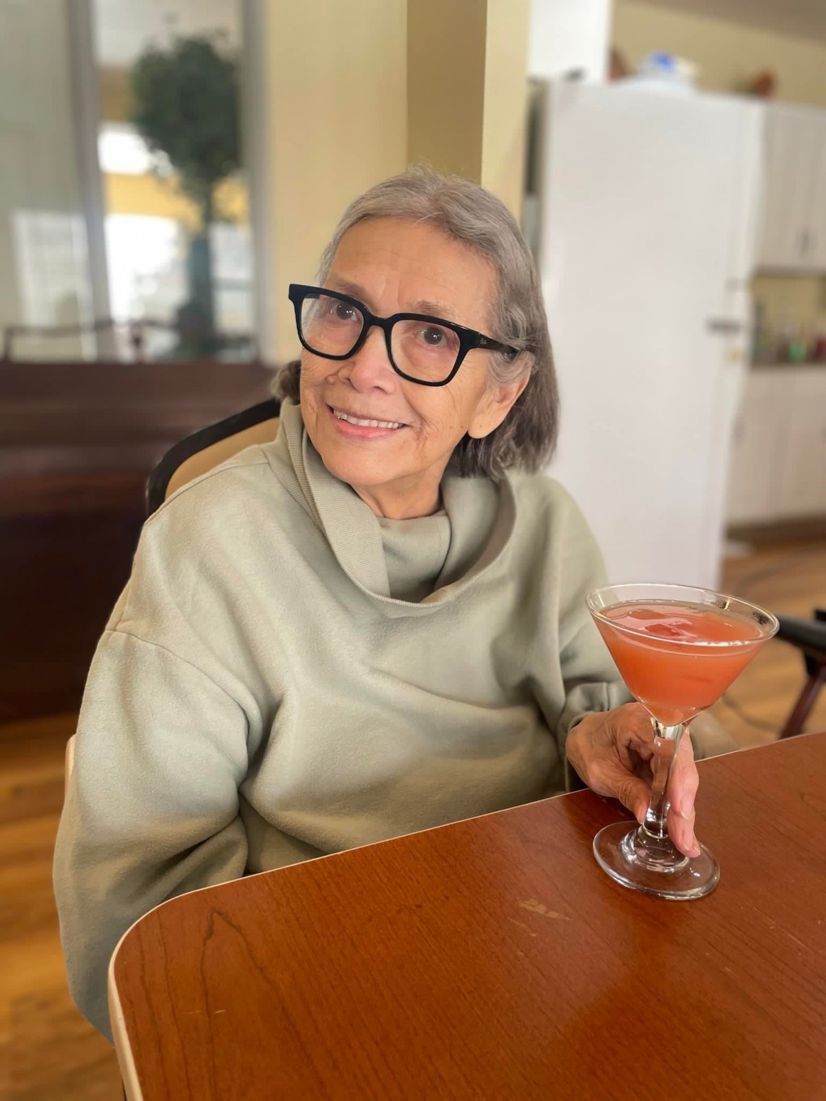 An elderly woman with glasses and gray hair smiles while holding a drink indoors.
