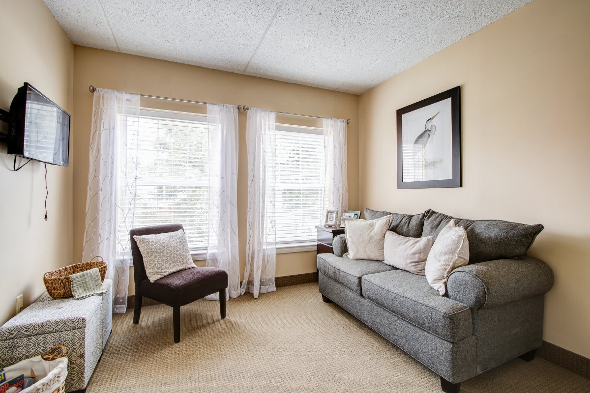 A cozy living room with a gray sofa, cushions, and a chair, illuminated by natural light through two large windows.