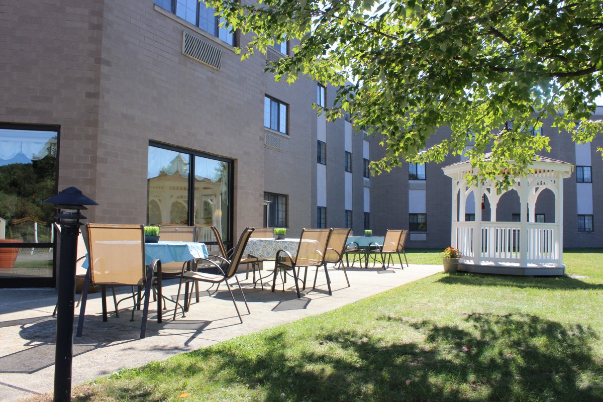 A sunny outdoor patio area with chairs and tables next to a building, featuring a gazebo on a well-maintained lawn.