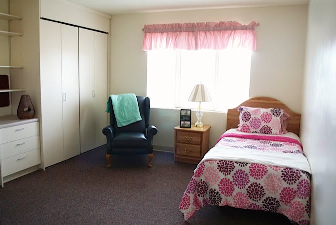 A cozy bedroom with a floral-patterned bedspread, a comfortable chair, and a window with pink curtains.