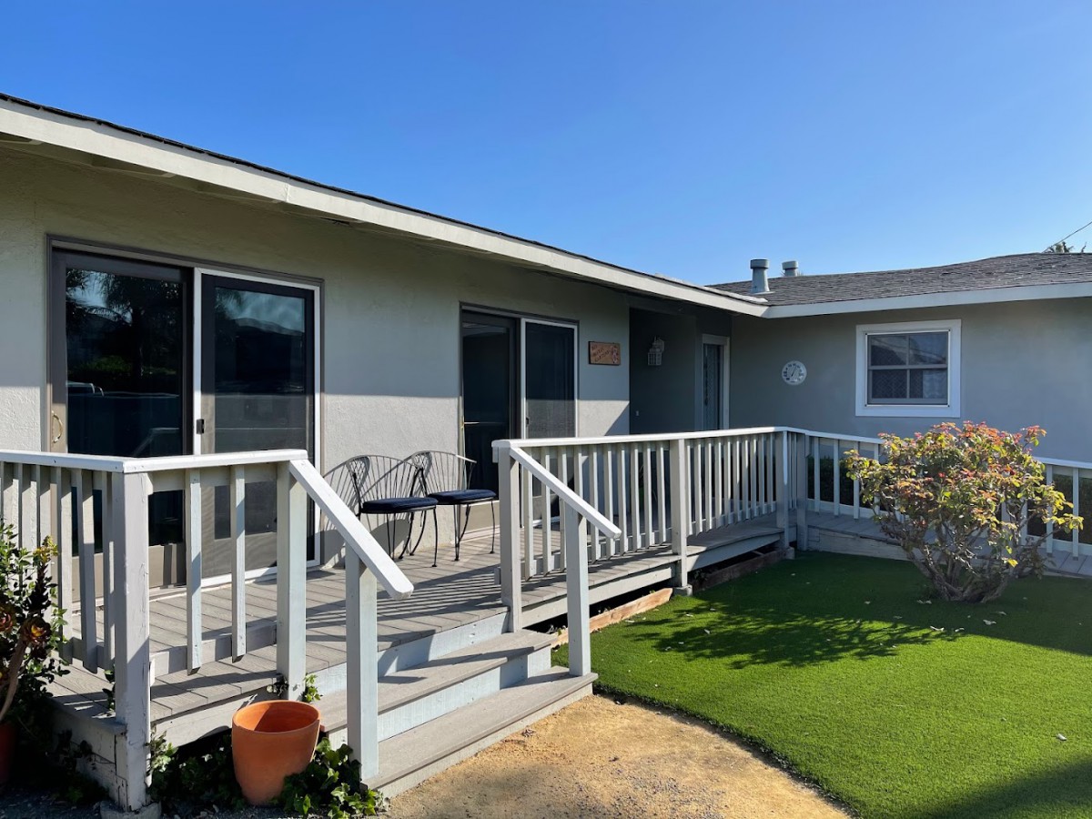 Sunny day view of a house with a wooden deck and railing, featuring outdoor furniture and a garden area.