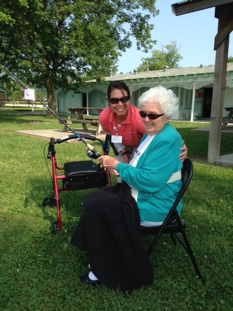 An elderly woman sits outdoors in a park with a caregiver, smiling and enjoying the sunny day.