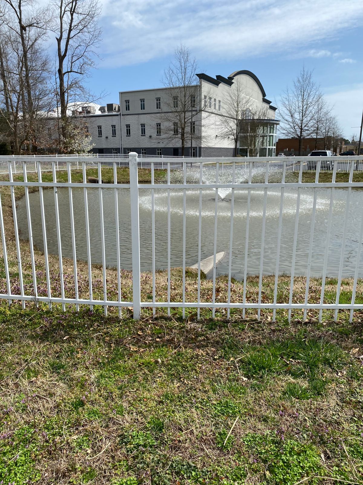 A fenced pond with a central fountain in front of a large building.