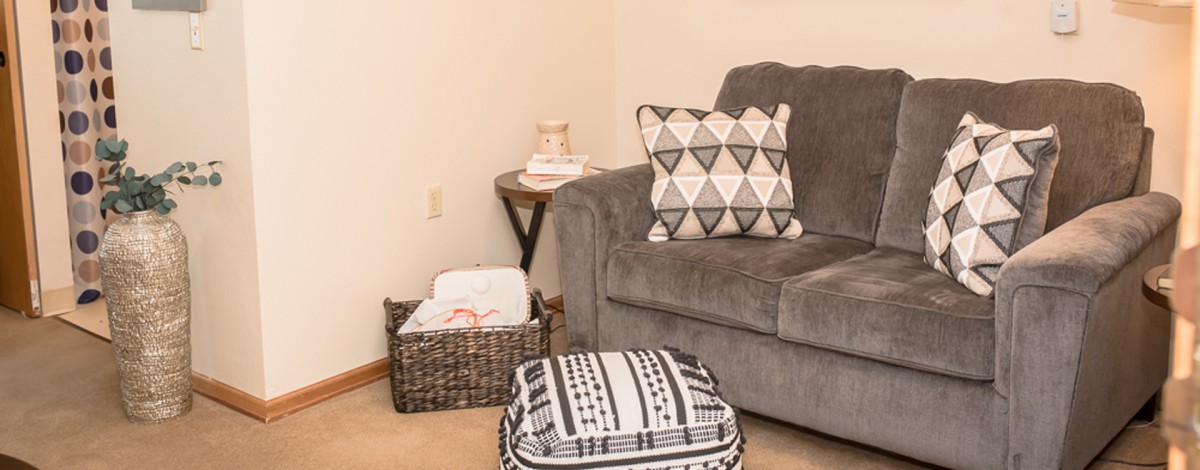 A cozy corner of a living room featuring a gray sofa with patterned cushions, a woven basket, and a geometric ottoman.
