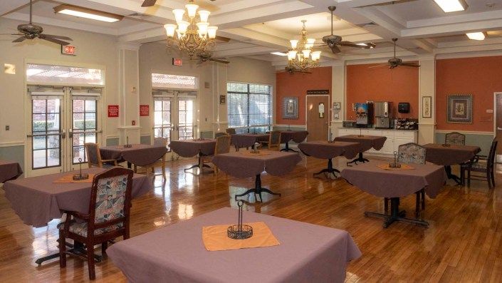 A spacious dining area featuring neatly arranged tables with purple tablecloths, elegant chandeliers, and wooden flooring.