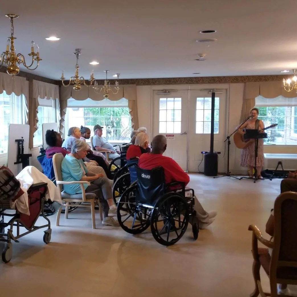 A group of elderly individuals in a nursing home setting, seated and listening attentively.