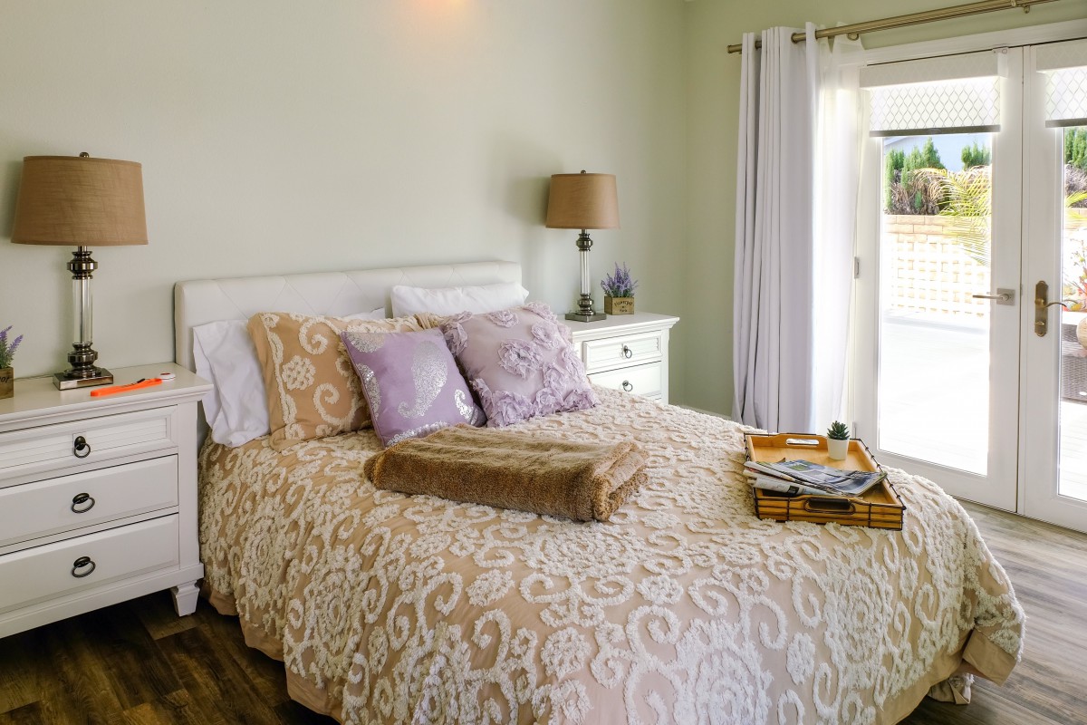 A cozy bedroom featuring a comfortable bed with patterned bedding, soft pillows, a bedside lamp, and natural light streaming through large windows.
