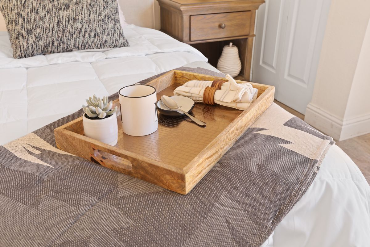 A cozy bedroom setting featuring a wooden tray with a mug, succulent, and napkins on a bed.