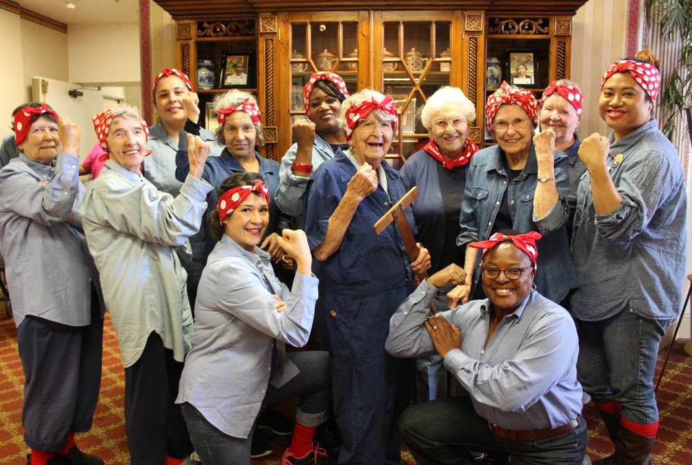 A group of women dressed as Rosie the Riveter, posing with red bandanas and fists raised in a celebratory gesture.