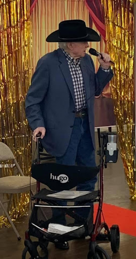 A man in formal attire is standing by a wheelchair, holding a cane and smiling at the camera against a shiny gold backdrop.