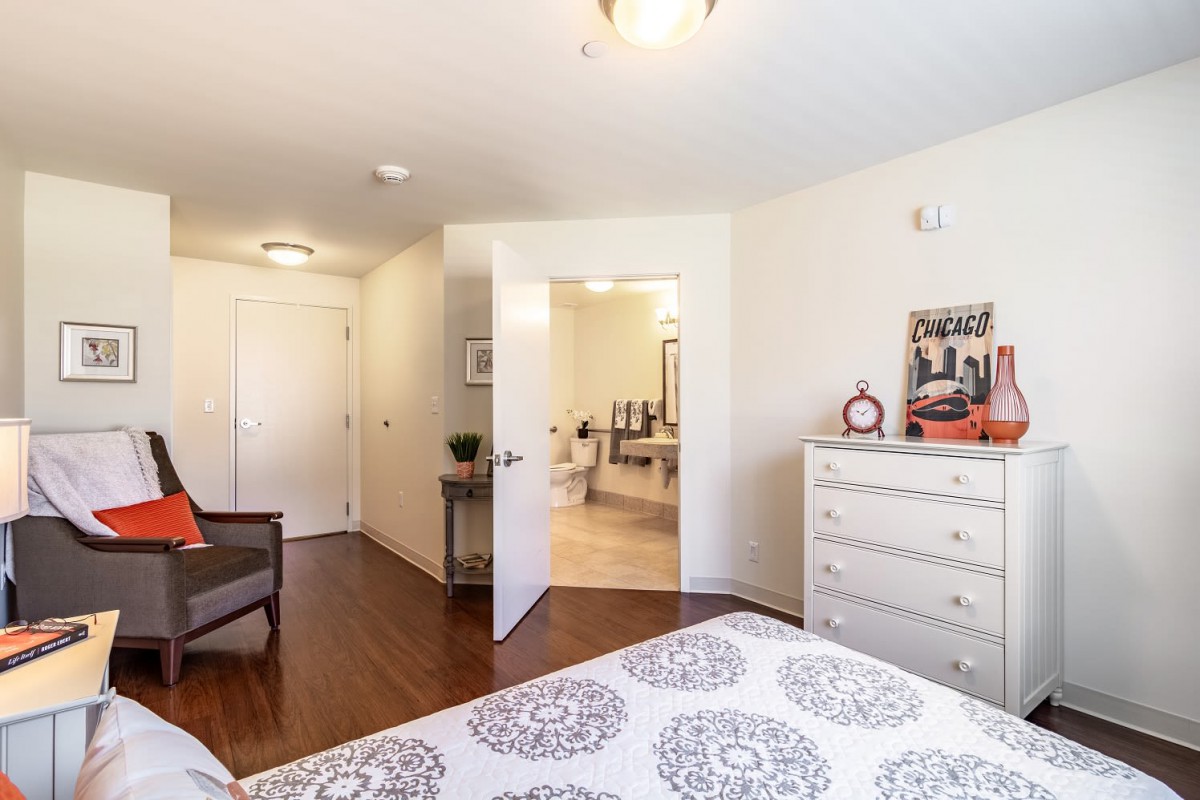 A cozy bedroom with wooden flooring, a neatly made bed, and a dresser, leading into a bathroom.