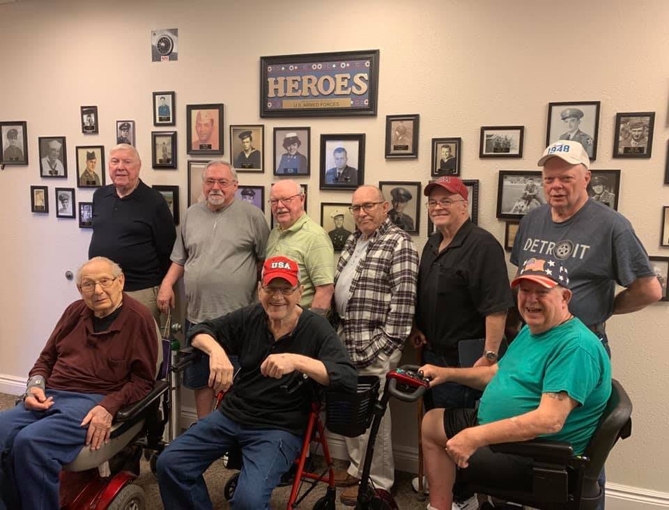 A group of seniors, some in wheelchairs, gather for a photo surrounded by framed photographs.