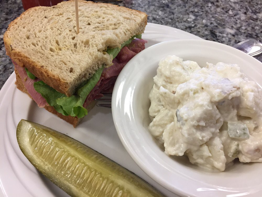 A classic deli sandwich with lettuce and meat, served alongside a dill pickle spear and a bowl of potato salad.
