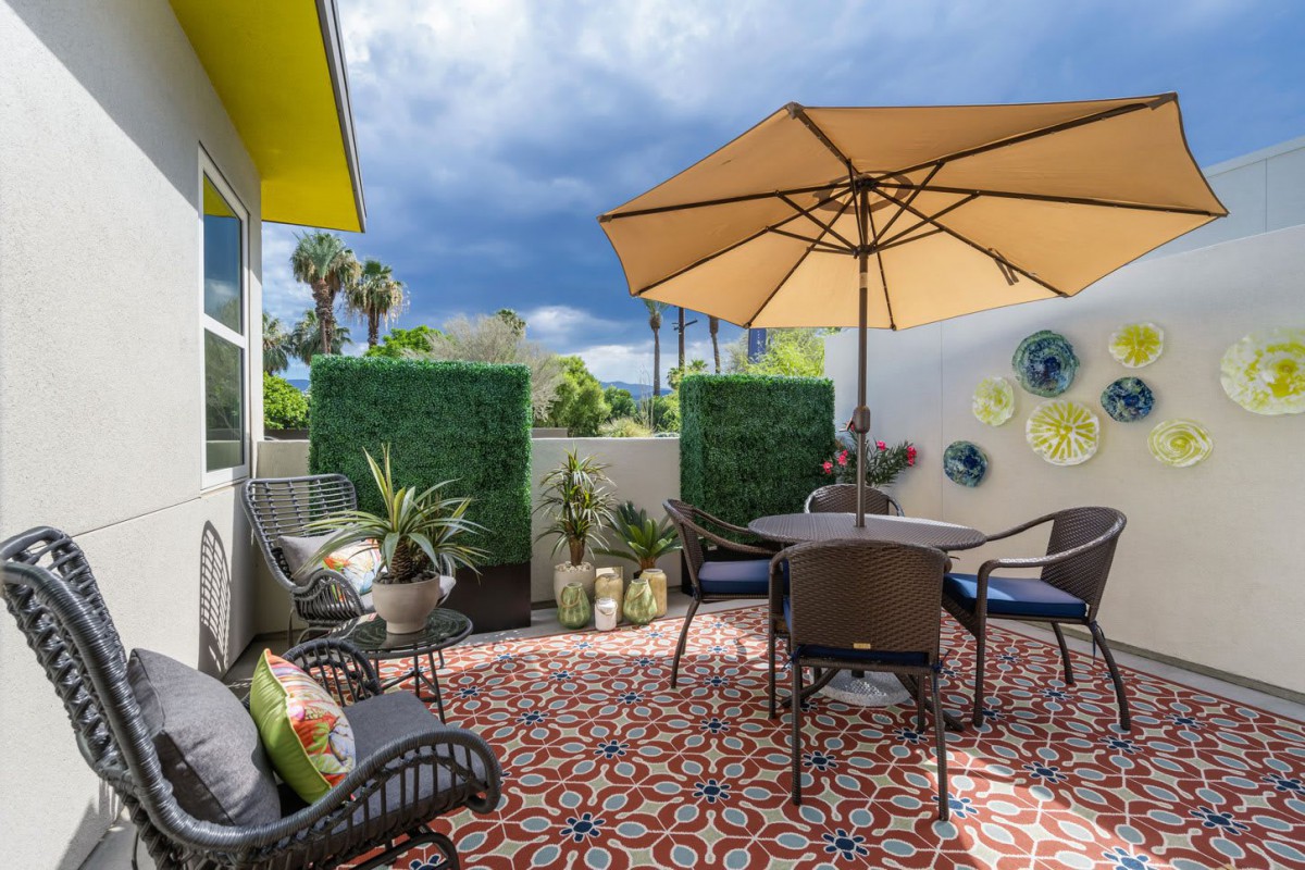 A cozy patio with outdoor furniture, a large umbrella, and vibrant decorative tiles surrounded by lush green plants.